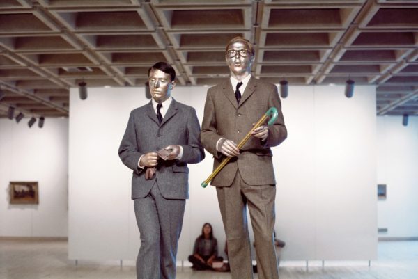 Two men dressed in elegant tweed suits and ties, with serious expressions, and their skin covered in metallic paint, stand on a table in a gallery. One man holds a cane, and the other holds a glove. In the background, three young people watch the men's performance.