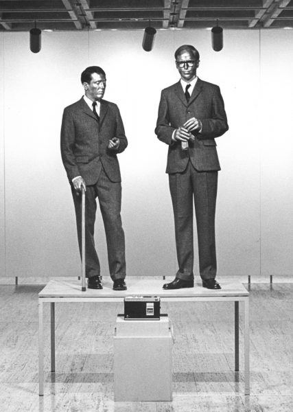 Two men dressed in elegant tweed suits and ties, with serious expressions, and their skin covered in metallic paint, stand on a table in a gallery. One man holds a cane, and the other holds a glove. An audio cassette player sits on a plain white plinth.