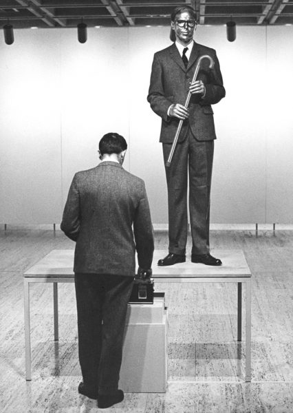 Two men dressed in elegant tweed suits and ties, with serious expressions, and their skin covered in metallic paint. One man stands on a table, holding a cane. The other is switching on an audio cassette player.