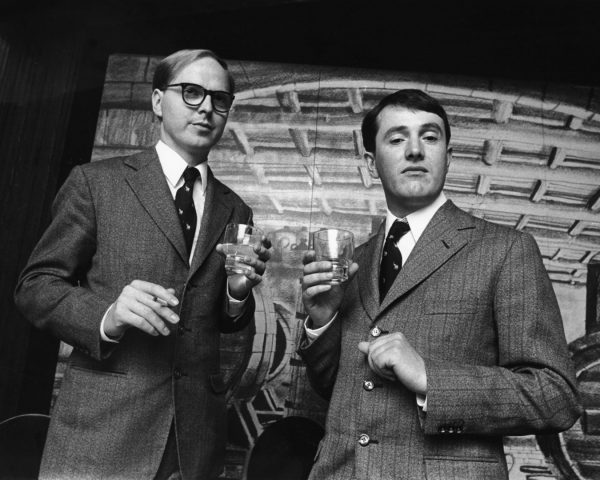 Two men wearing elegant tweed suits and ties, holding cocktail glasses and a cigarette, with amused expressions.
