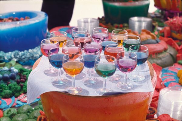 Bright multi-coloured food, arranged carefully on platters on a tiered table. In the foreground is a tray of wineglasses, holding drinks in various bright colours.