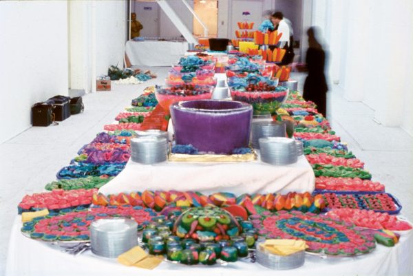 Bright multi-coloured food, arranged carefully on platters on a tiered table, with a moulded bowl of purple-coloured ice, and an arrangement of fruit encased in jelly. Four or five people, dressed in black and white, are busily moving near the table.