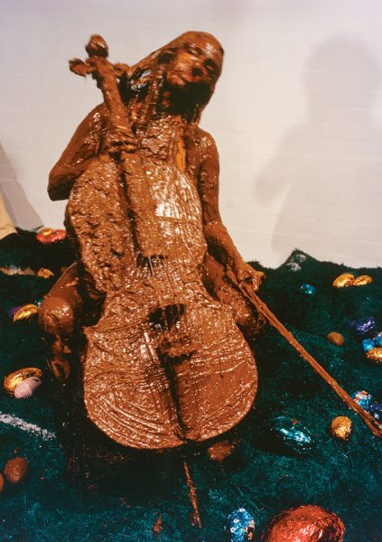 A naked woman, seated, holding a cello and cellist’s bow. Both the woman and cello are completely smothered in chocolate. The floor is covered with crumpled artificial turf, with a scattering of large, colourful easter eggs.