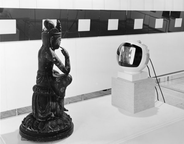 A small statue of a seated Buddha faces towards a 1960s style, spherical television monitor. On the television screen is a video image of the Buddha statue.