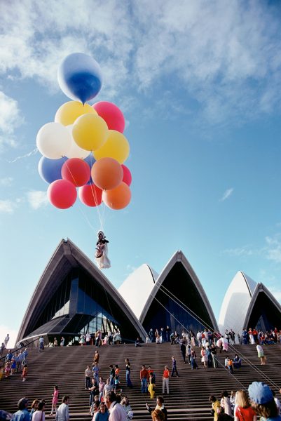 A woman suspended by a bunch of large, brightly coloured balloons, high above the forecourt of the Sydney Opera House, against a bright blue sky. The woman plays a cello, wearing a white skirt and headdress with black leotard. A scattered crowd of people watches the woman’s performance.