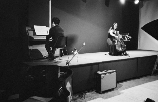 A man and woman, seated on a small, bare stage. The woman plays a cello, dressed in a simple black evening gown. The man wears a black suit and plays an upright piano. In front of the stage are a microphone stand, 1970s style amplifier, grand piano and electrical cables.