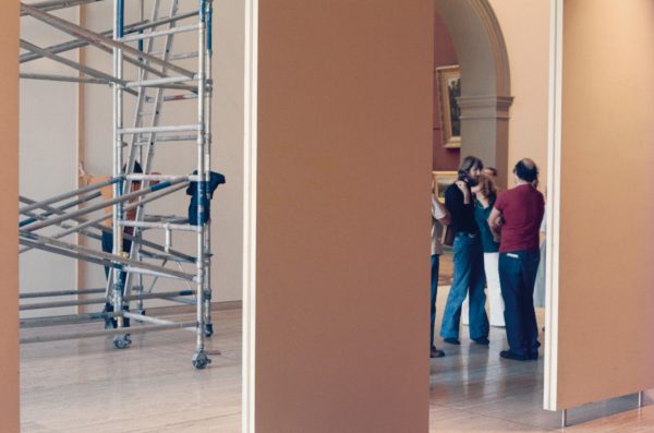 A small group of people gathered in conversation in the Art Gallery of New South Wales entrance court. One man leans on scaffolding and holds a long measuring stick against the gallery wall.