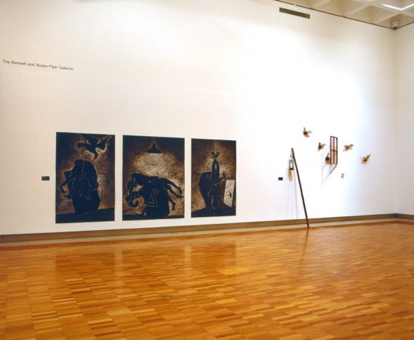 Three large charcoal drawings of male and female figures contorted and suspended upside-down in a darkened room, with a swooping black crow. The drawings are mounted on the wall of a gallery, alongside an installation of objects, including a tall stick, glass lamp, window frame and small flying creatures.