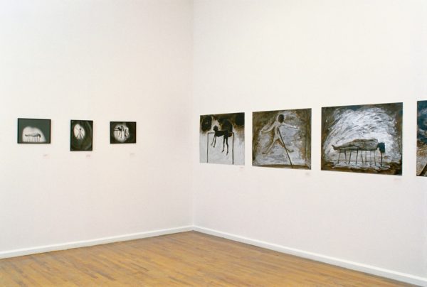 A series of black-and-white photographs and small paintings of a female figure, contorted and suspended by long sticks, lit by a spotlight. The artworks are mounted on the wall of a gallery.