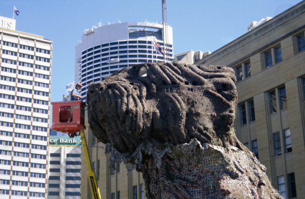 The head of a giant metal sculpture of a terrier puppy, covered in wire mesh. A man in a cherry picker is adjusting the mesh.