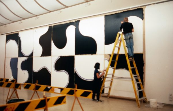 A young man and young woman paint a geometric arrangement of black and white arcs and curves across a long gallery wall. The man stands on a tall ladder.