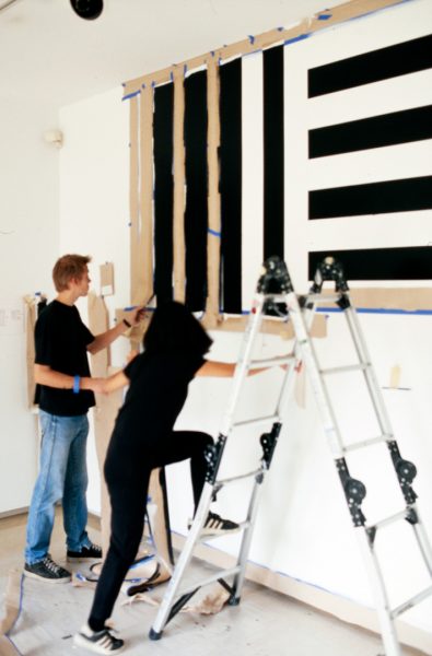 A young man and young woman paint a geometric arrangement of black and white bands across a gallery wall, marked out with masking tape and paper.