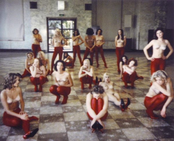 A group of 16 young, slim white women, dressed identically in bras, red tights and red stiletto shoes, stand and crouch in the marble hall of an art gallery. One woman is completely nude.
