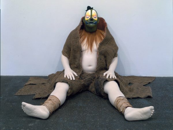 A life-sized mannequin of a clown sits, eyes closed, against the wall of a contemporary art gallery. The clown is wearing dark green face paint, a tiny hat, rough hessian clothing and a neckpiece made of hair, with their belly and legs exposed.