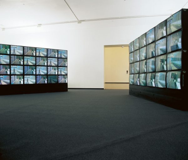 Two large banks of 24 video monitors, stacked in a contemporary art gallery. Each monitor displays the video image of a man and woman walking alone in a city.
