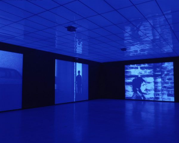 Three large blue-and-white video projections on the walls of an art gallery, which cast blue light across the darkened room. The video images feature a man walking past a brick wall.