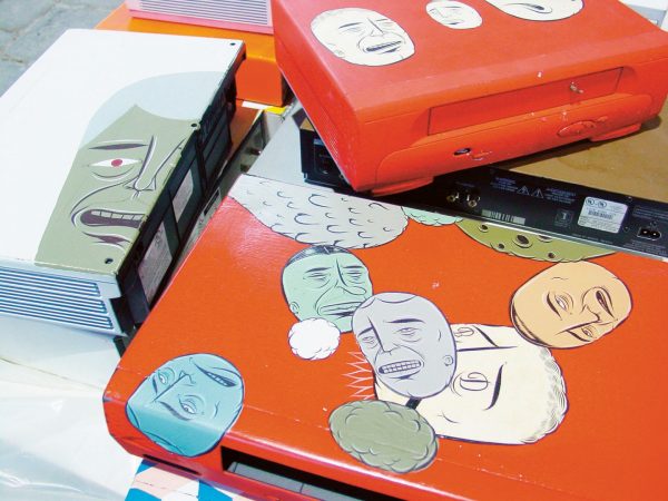 A pile of video cassette players, painted in orange, with the cartoon-like faces of grimacing men.