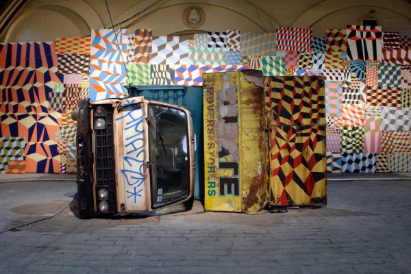 An overturned dump truck, graffitied with tags and geometric patterns. In the background is a wall of colourful, irregular geometric shapes in a checkerboard pattern.