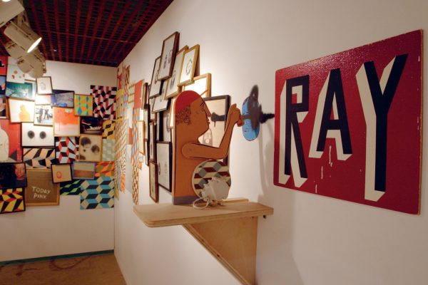 A simple, mechanical wooden structure, featuring the cartoon-like figure of a man holding a can of spray paint. In the background are framed photos, drawings and geometric patterns, hung on the wall as a dense and overlapping assemblage.