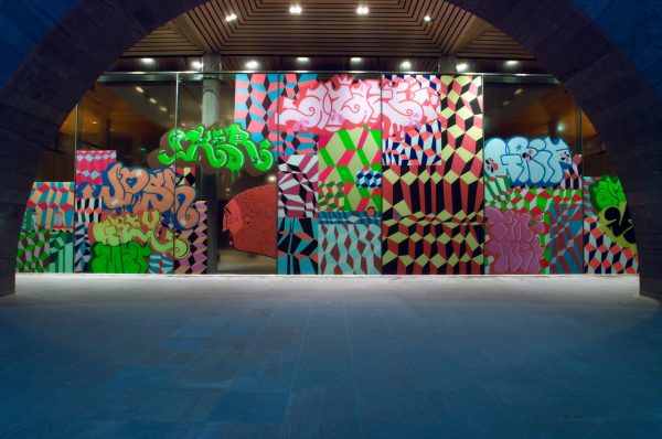 A glass wall covered in colourful, irregular geometric shapes, graffiti tags and the cartoon-like face of a weary old man, arranged in a checkerboard pattern.