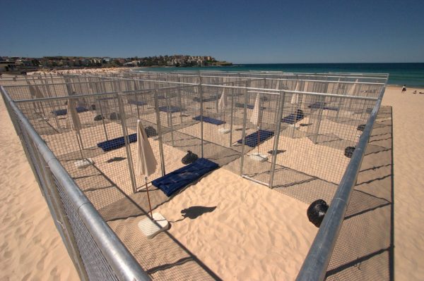 A grid of 21 cells, made of wire-mesh fencing, on the sands of Bondi Beach. Each cell contains a beach umbrella, air mattress and black plastic garbage bag.