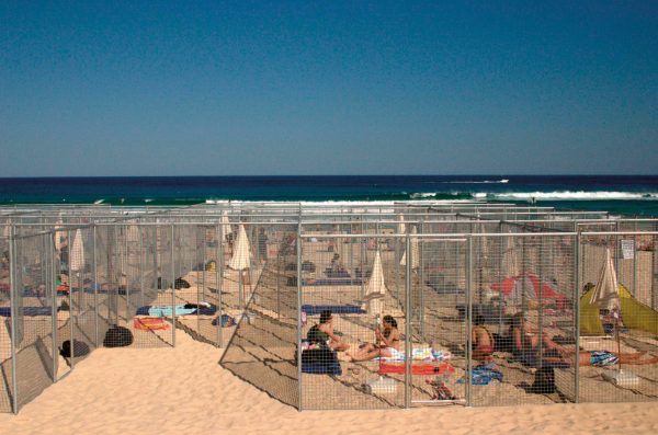 A grid of 21 cells, made of wire-mesh fencing, on the sands of Bondi Beach. Inside each cell are relaxing beachgoers, with a beach umbrella, air mattress and black plastic garbage bag.
