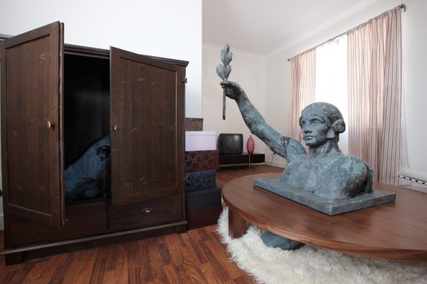 A modern domestic living room, with timber floor, shag pile rug, coffee table, plant and a wooden cupboard. On top of the coffee table is the head and shoulders of a large bronze statue of a woman, holding an olive branch. Inside the cupboard is the head of a bronze statue of a horse.