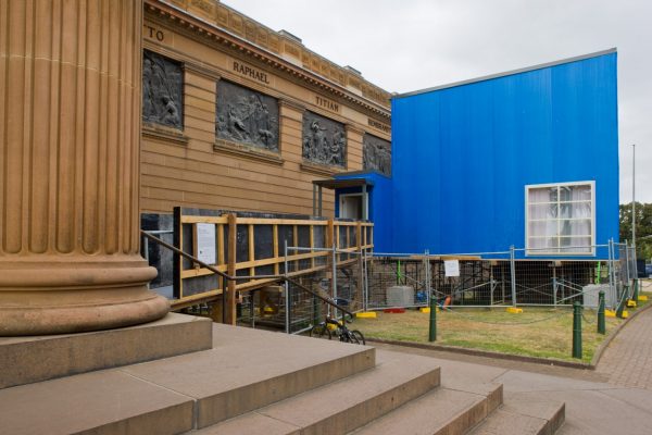 Close-up of a sandstone neo-classical museum. Next to the building is a small, blue structure with a window, on top of a scaffolding base, with scaffolding ramp.
