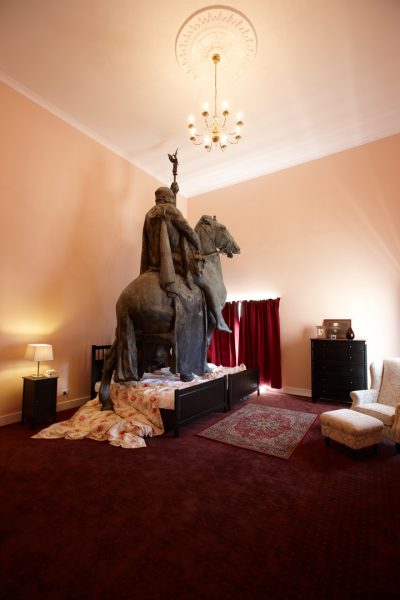 A tidy domestic bedroom with two single beds, furnished with deep red carpet, curtains, an armchair and a chandelier. On top of the beds is a large bronze statue of a man, wearing a cape and holding a staff, on horseback. The bed covering is crumpled under the horse’s hooves, and spilling onto the floor.
