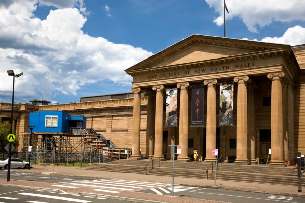 A sandstone neo-classical museum. Next to the building is a small, blue structure with a window, on top of a scaffolding base, with scaffolding stairs.