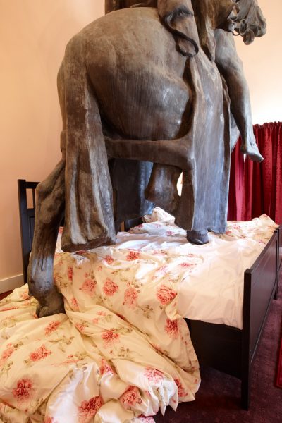 Close-up of a large bronze statue of a horse, climbing onto two single beds. The bed covering is crumpled under the horse’s hooves, and spilling onto the floor.