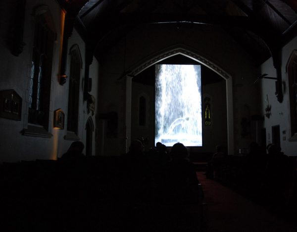 A video plays on a tall, narrow screen in a dark neo-Gothic church. The video shows a man in a white robe, levitating, in a gushing torrent of water.