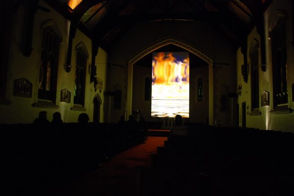 A video plays on a tall, narrow screen in a dark neo-Gothic church, with a small group of people seated, watching. The video shows a blazing fire reflected in a pool of water.