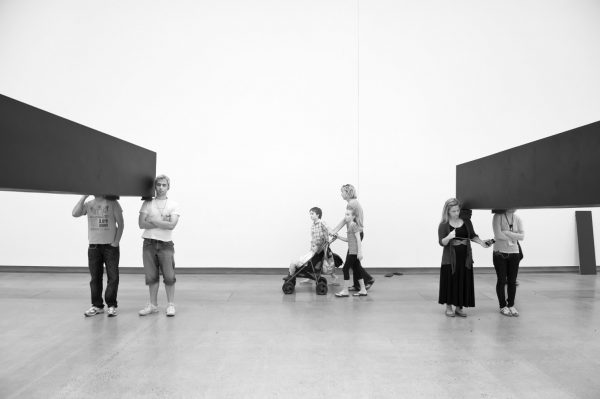 2 pairs of young people stand in line in an art gallery. Between each pair, carried on their shoulders, is a long, dark, heavy rectangular object. A woman with a stroller and three children walks past.