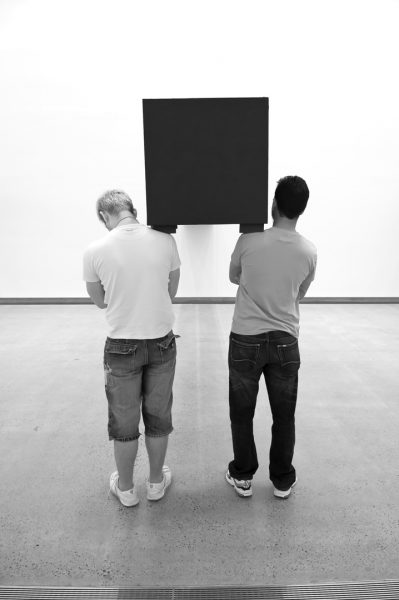 Two young men in an art gallery, facing towards the wall. Between the men, carried on their shoulders, is a long, dark, heavy rectangular object.
