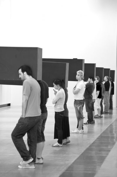 Aerial view of 7 pairs of people stand in line in an empty art gallery. Between each pair, carried on their shoulders, is a long, dark, heavy rectangular object.
