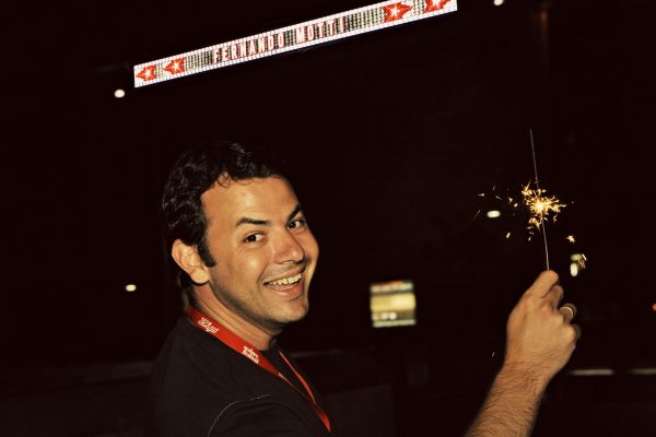 A young man holds a glowing sparkler. In the background, the name Fernando Motti is written in bright lights, with a pattern of red and yellow stars, against a dark night sky.