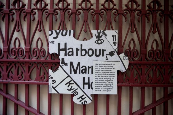 A large white puzzle piece, showing the fragment of a street map, hangs on an ornate metal fence. On the map is written a short story, which reads, “My work driveway is on Pitt Street and getting out at peak hour can be tough, as commuters rush to Wynyard Station to head home at the end of the day. Most nights I rely on an everyday act of kindness to get out of the driveway – a busy commuter stopping at the drive and ushering me on so I can get my car out. Thank you!”