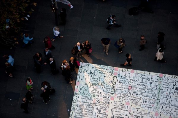 A large, hand-drawn street map of the Sydney CBD area, installed on the paving of a large, open outdoor space. A small crowd of people of around 40 people is gathered around the map.