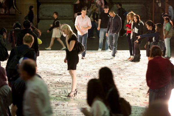 A small, diverse crowd of people walks across a large, hand-drawn street map, in an open outdoor space, during the evening. The people are pointing and looking down intently at the map. Some are dressed in business clothes, some in casual attire.