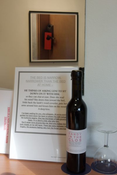 A bottle of wine, a plain wine glass, and pages of laminated, written notes propped against a mirror. The mirror reflects a framed photograph on the opposite wall. The photograph shows the detail of a modern hotel door, with a plain red sign hung on the handle.