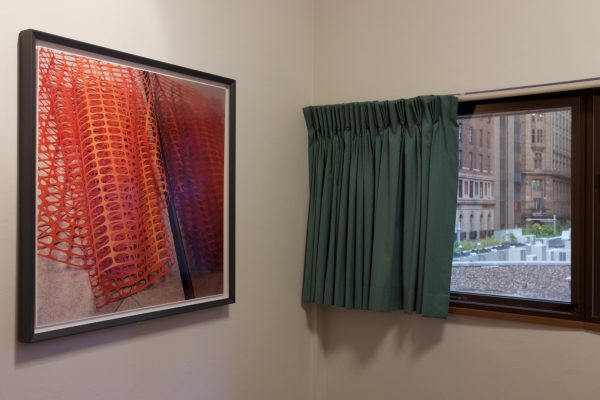A simple room with white walls, a window with plain curtains, looking onto city buildings, and a framed photograph on the wall. The photograph shows a sheet of orange plastic mesh, piled on a paved floor.