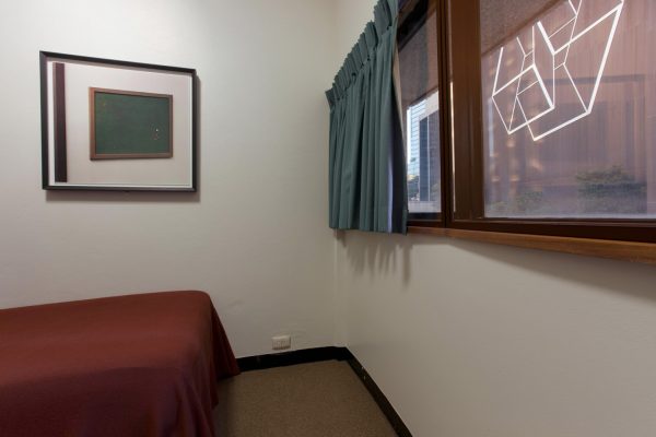 A simple hotel room with white walls, a single bed with a plain bedspread, a window looking onto city buildings, and a framed photograph on the wall. The photograph shows a green felt pinboard, with a small number of coloured pins.
