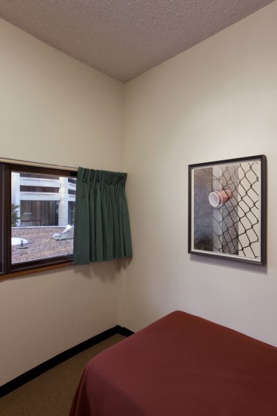 A simple hotel room with white walls, a single bed with a plain bedspread, a window looking onto city buildings, and a framed photograph on the wall. The photograph shows two disposable cups wedged into a chain link fence.