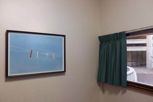 A simple room with white walls, a window with plain curtains, looking onto city buildings, and a framed photograph on the wall. The photograph shows four coloured pegs on a twisted clothes line.