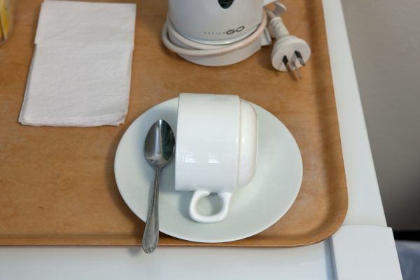 The detail of a plain white coffee cup, on its side, with a teaspoon, on a plain white saucer. The cup and saucer sit on a wooden tray, with plain white napkins, and a small electrical appliance.