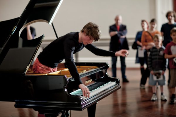 A young man stands inside a grand piano, and leans forward to play the keys with a look of intense concentration. The piano has a round hole cut in the lid. Around 7 people are watching the pianist, inside a gallery room.
