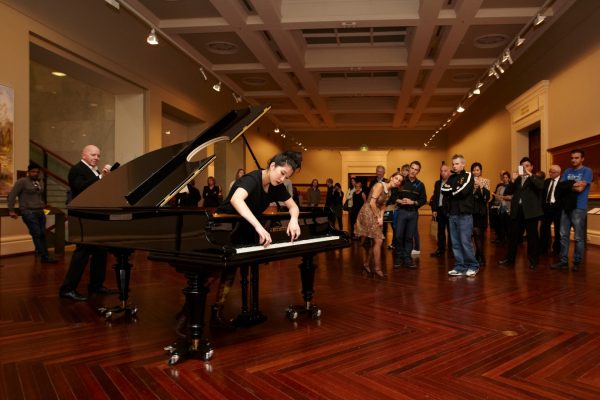 A young woman stands inside a grand piano, and leans forward to play the piano keys, with a look of intense concentration. The piano has a round hole cut in the lid, and wheels attached to the legs. A small crowd of around 30 people is watching the pianist, inside a long, ornate gallery.