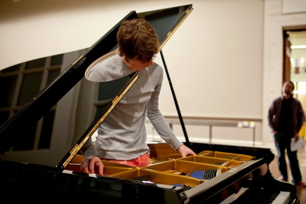 A young man stands inside a grand piano, and strums the piano strings, with a look of intense concentration. His head emerges through a round hole, cut into the lid of the piano.