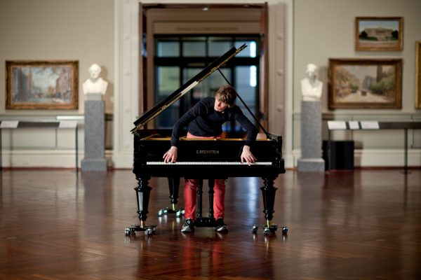 A young man stands inside a grand piano, and leans forward to play the keys with a look of intense concentration. The piano has a round hole cut in the lid, wheels attached to the legs, and the name C Bechstein in gold lettering on the front. Behind the piano is an ornate gallery with gilt-framed pictures and marble busts on plinths.